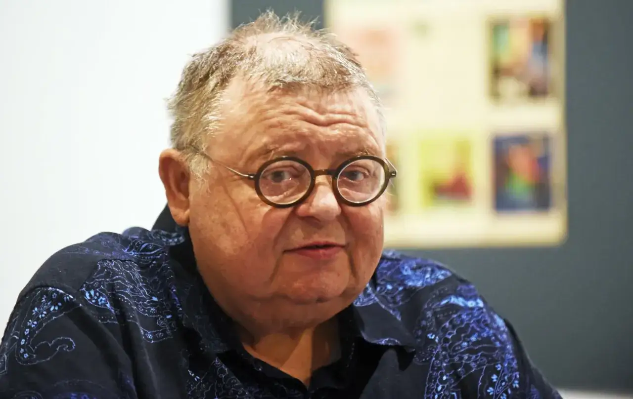 Wojciech Mann w okularach, z siwiejącymi włosami i w granatowej koszuli z wzorem. Wiek nie odbiera mu charyzmy.