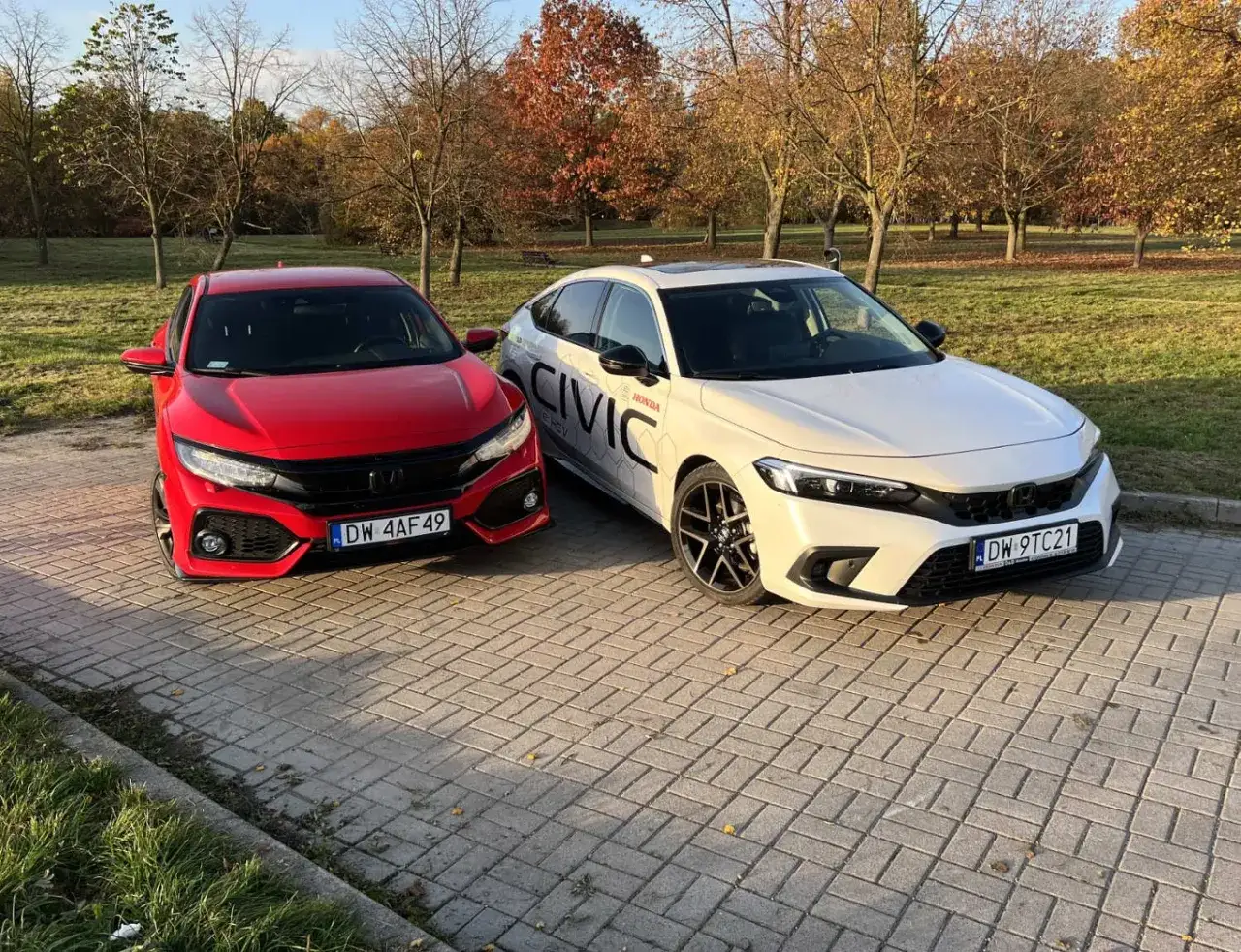 Dwa samochody Honda Civic 10: czerwony sportowy i biały sedan, zaparkowane na tle jesiennego parku.