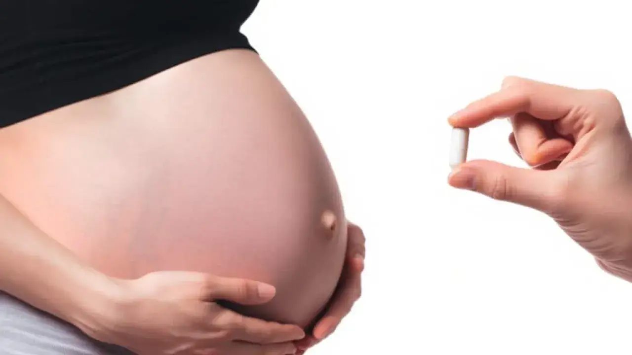Dłoń trzyma białą kapsułkę Actiferol obok brzucha kobiety w ciąży.