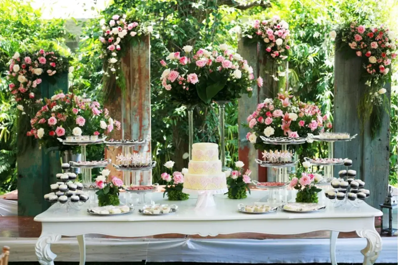 Mesa de dulces con pastel de tres pisos, cupcakes y postres variados, decorada con flores rosas y blancas. Ideal para hacer flores de golosinas.
