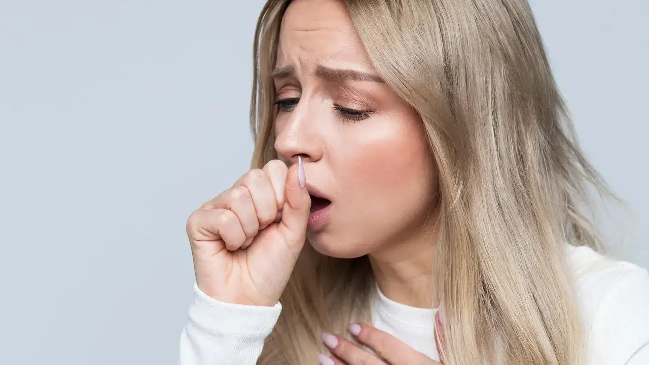 Mujer rubia con expresión de malestar tose, llevándose la mano a la boca. Siente dolor en el pecho al toser sin fiebre.