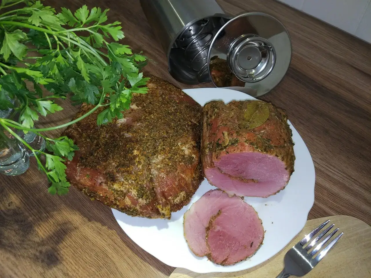 Gotowy do parzenia schab po wędzeniu, przyprawiony ziołami, z liściem laurowym. Obok świeża natka pietruszki i wędzarka.
