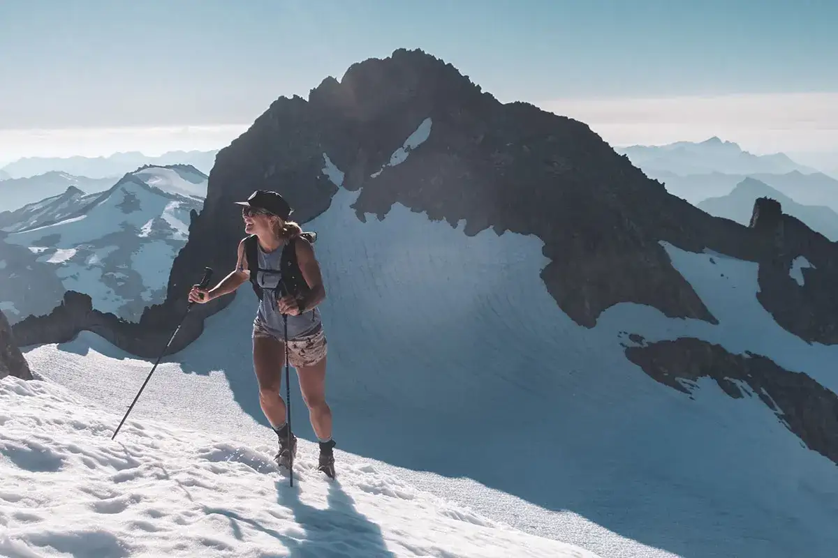 Frau beim Skyrunning auf schneebedecktem Berggipfel, mit Stöcken und Rucksack, im Hintergrund weitere Berge.