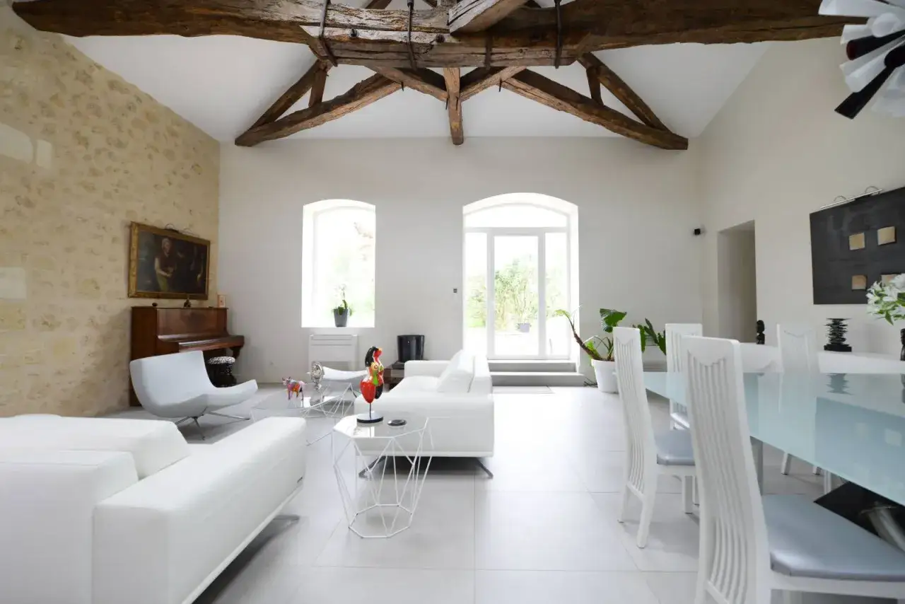 Avant après idée rénovation maison ancienne : salon lumineux avec poutres apparentes, mur en pierre, piano, mobilier blanc moderne et salle à manger.