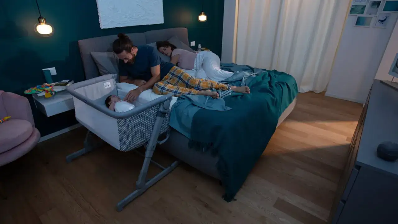 Ein Vater wacht &uuml;ber sein Neugeborenes im Beistellbett, w&auml;hrend die Eltern im Bett schlafen. Die Frage, wie viel schlafen Neugeborene, besch&auml;ftigt viele Eltern.