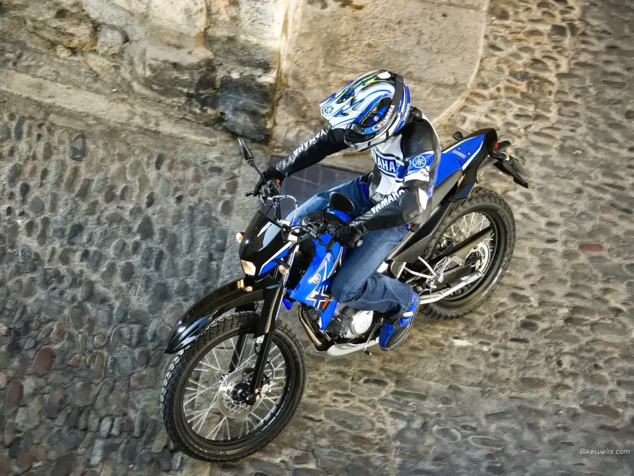 Jaki motocykl enduro! Kierowca w kasku i kurtce Yamaha przemierza brukowaną uliczkę na niebieskim motocyklu.
