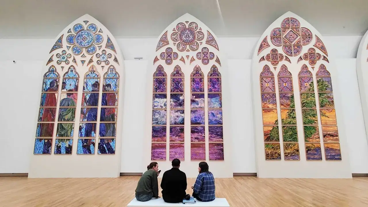 Trois vitraux modernes, inspir&eacute;s de l'art parisien, ornent un mur blanc. Trois visiteurs contemplent ces &oelig;uvres color&eacute;es.