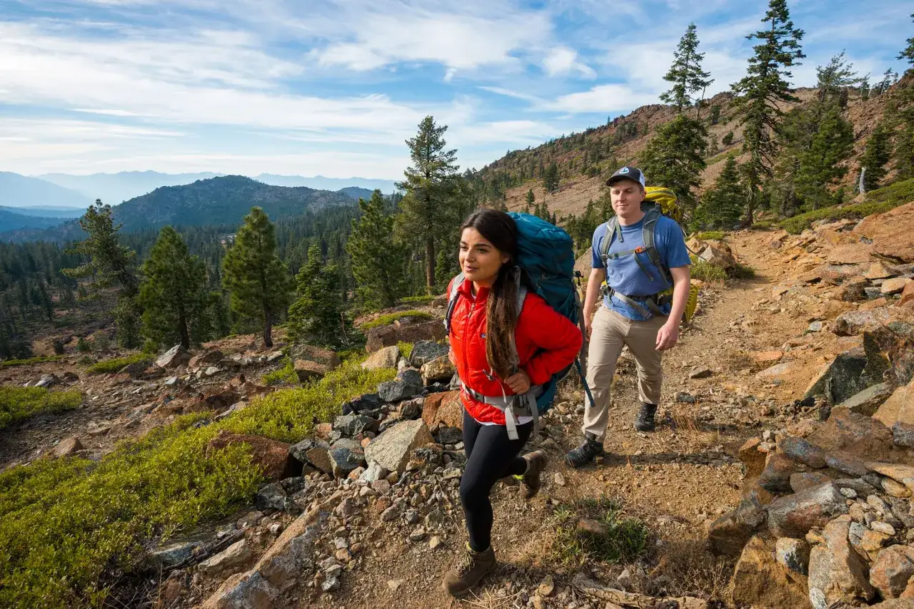 Två vandrare med stora ryggsäckar går på en stenig stig. De njuter av utsikten över bergen och skogen längs Pacific Crest Trail.