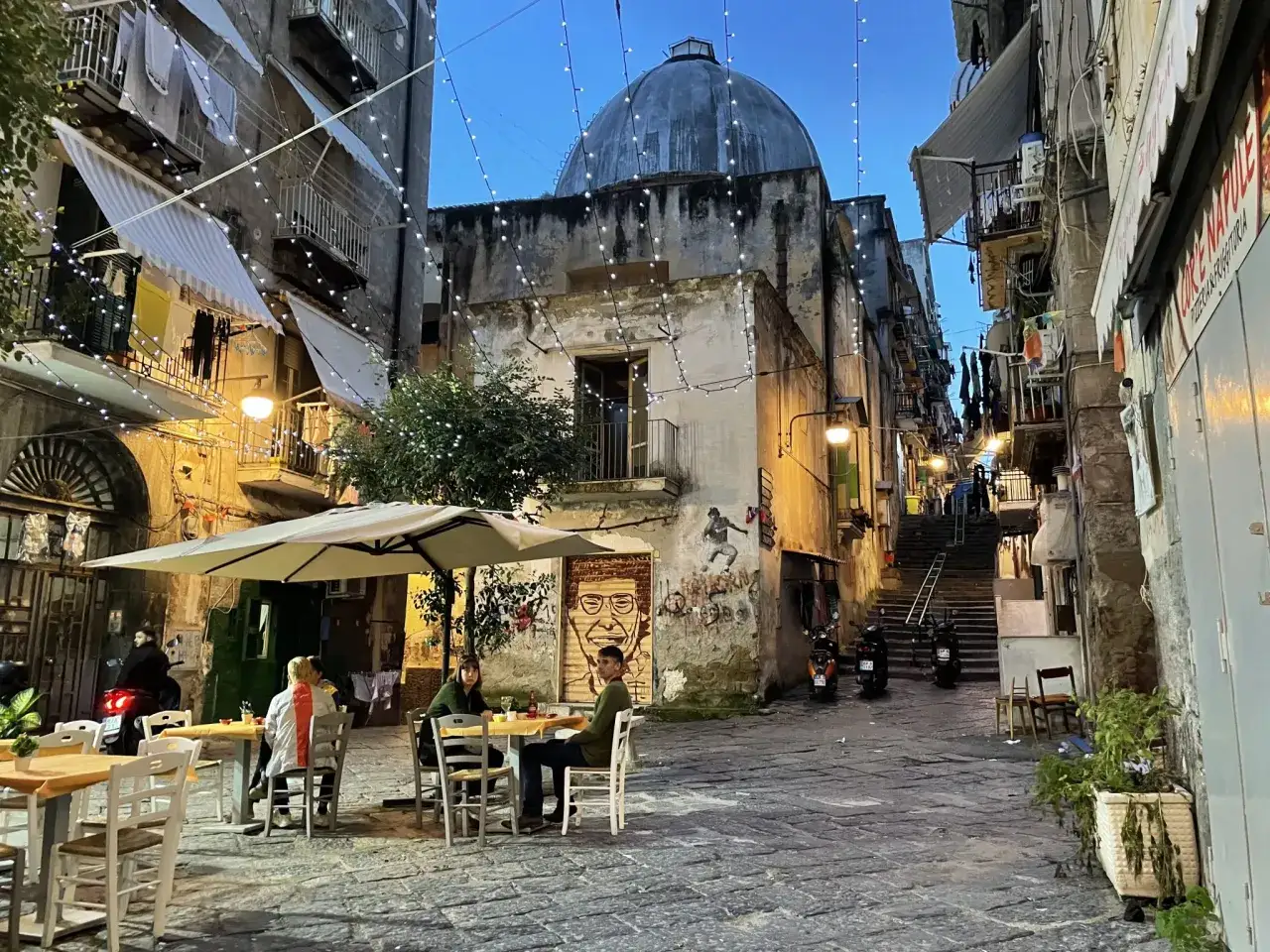 Enjoying a meal at an outdoor cafe in a charming Naples alleyway, with string lights and a dome in the background.