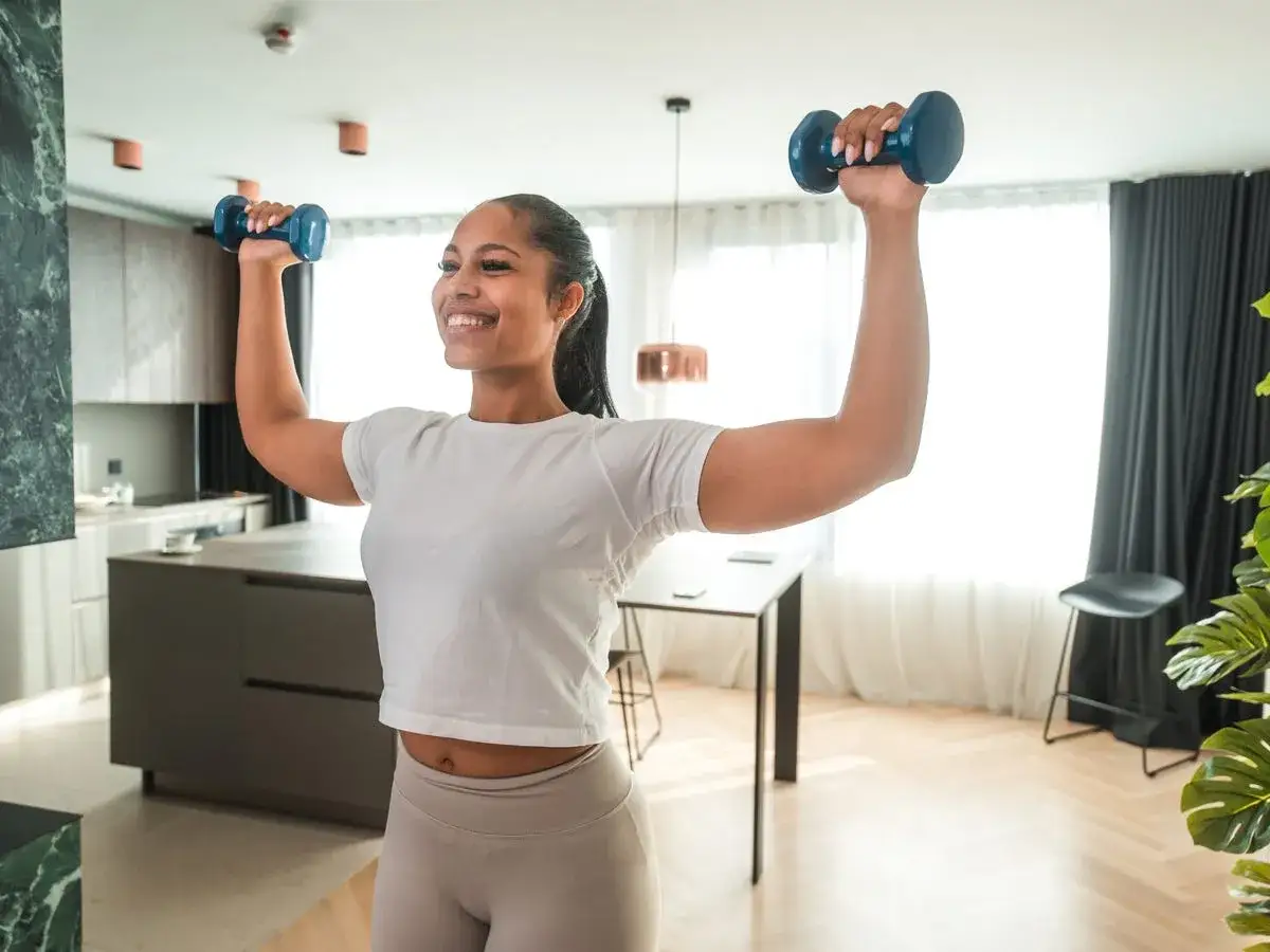 Femme souriante faisant du renforcement musculaire &agrave; domicile avec des halt&egrave;res bleus. Elle est en tenue de sport dans un appartement moderne.