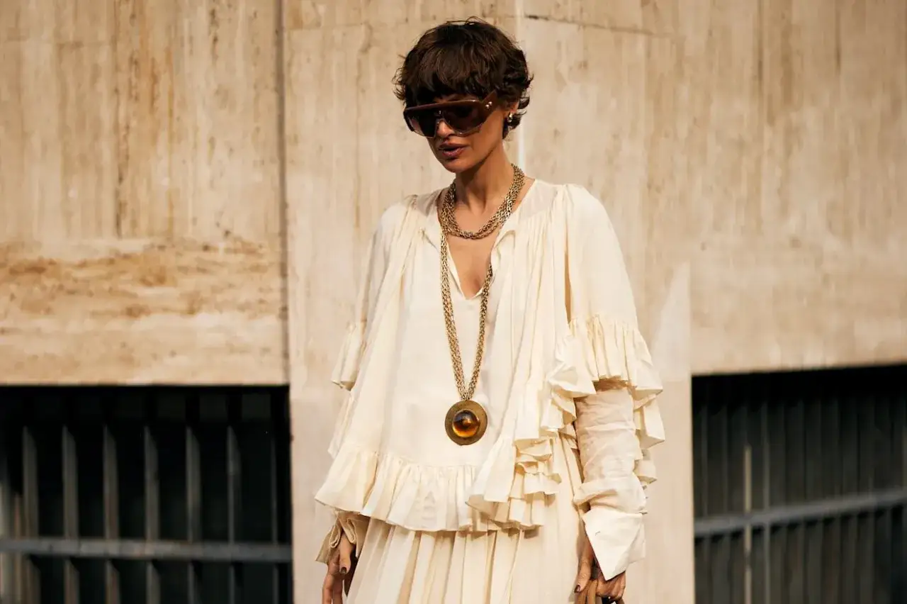 Mujer con vestido blanco voluminoso y volantes, collar dorado largo con medallón, gafas de sol y pelo corto. Un look que evoca el estilo boho.