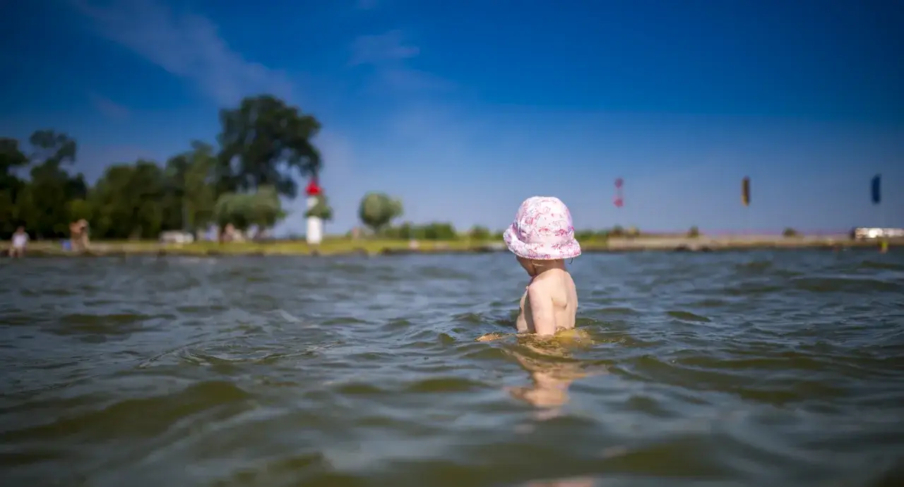 Kleines Kind mit rosa Hut planscht im Wasser am Stettiner Haff Strand. Im Hintergrund Bäume und ein Leuchtturm.