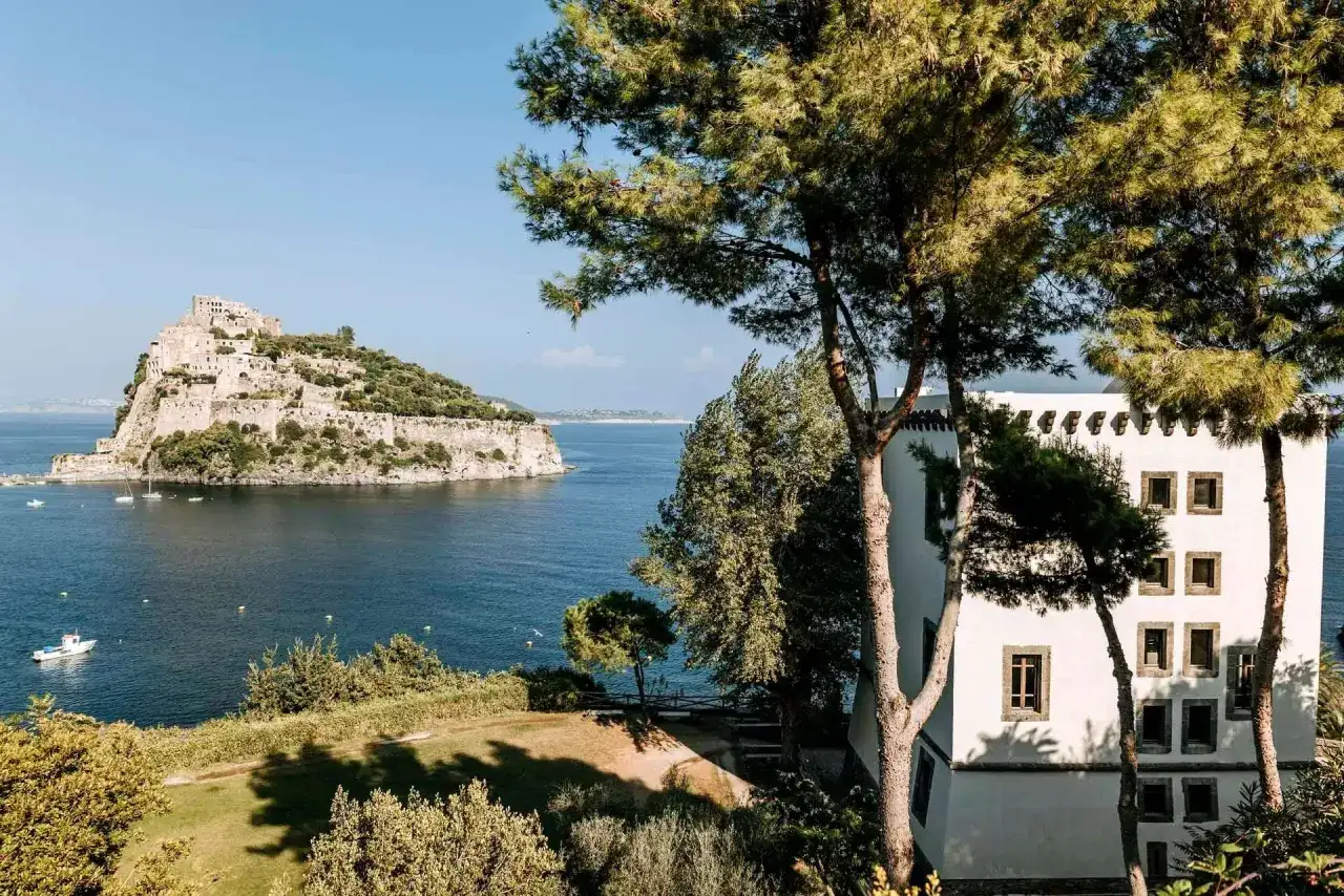 Vista del mare blu con la torre di Michelangelo a Ischia in lontananza, alberi verdi e una casa bianca in primo piano.