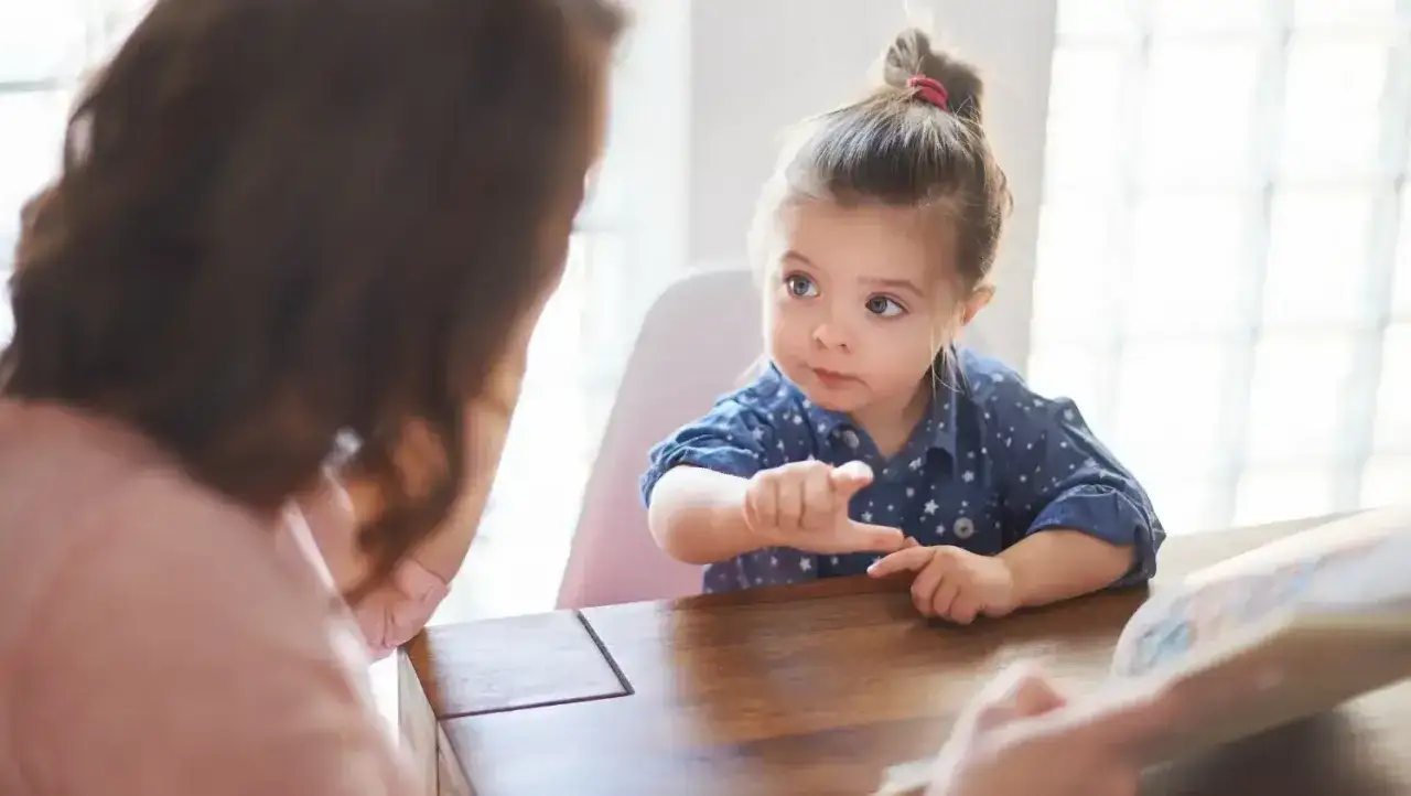 Mała dziewczynka z kucykiem, która nie radzi sobie z emocjami, wskazuje palcem na książkę, podczas gdy mama próbuje ją uspokoić.