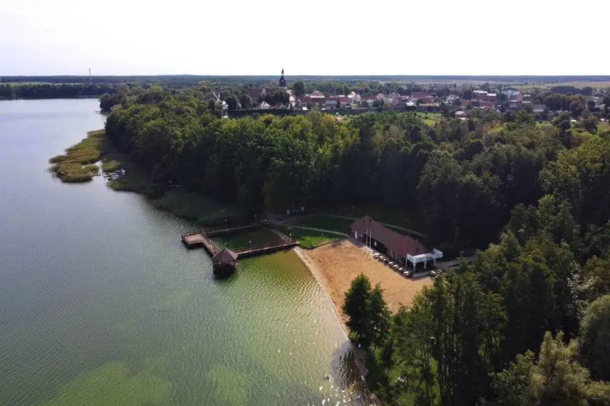 Widok na jezioro z pomostem i plażą, obok gardna i sarbska, w tle miasteczko z wieżą kościelną.