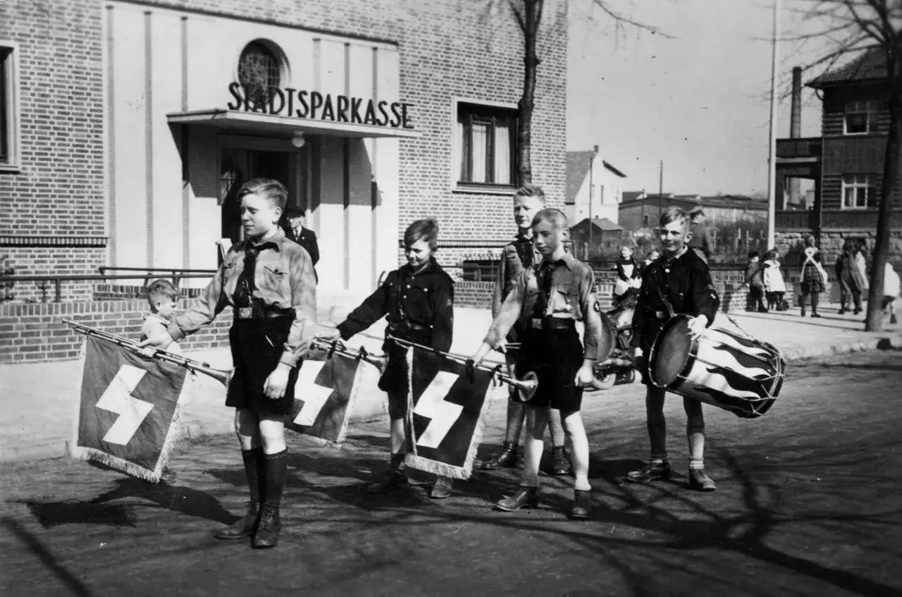 Jungen in Uniformen tragen Fahnen mit Hakenkreuzen und marschieren vor der Stadtsparkasse. Ein Schlagzeug ist ebenfalls zu sehen. Dies ist ein Einblick in den Alltag im Nationalsozialismus.
