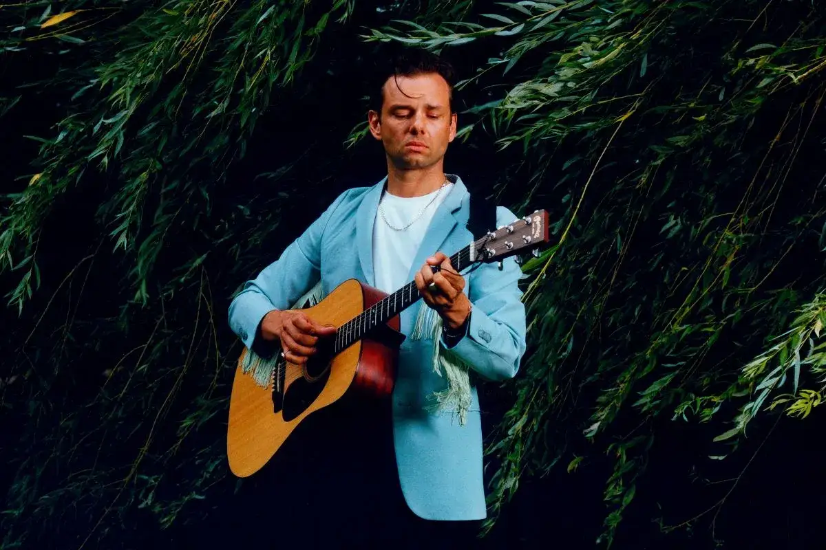 Rubens piosenkarz w błękitnej marynarce gra na gitarze wśród zielonych gałęzi.