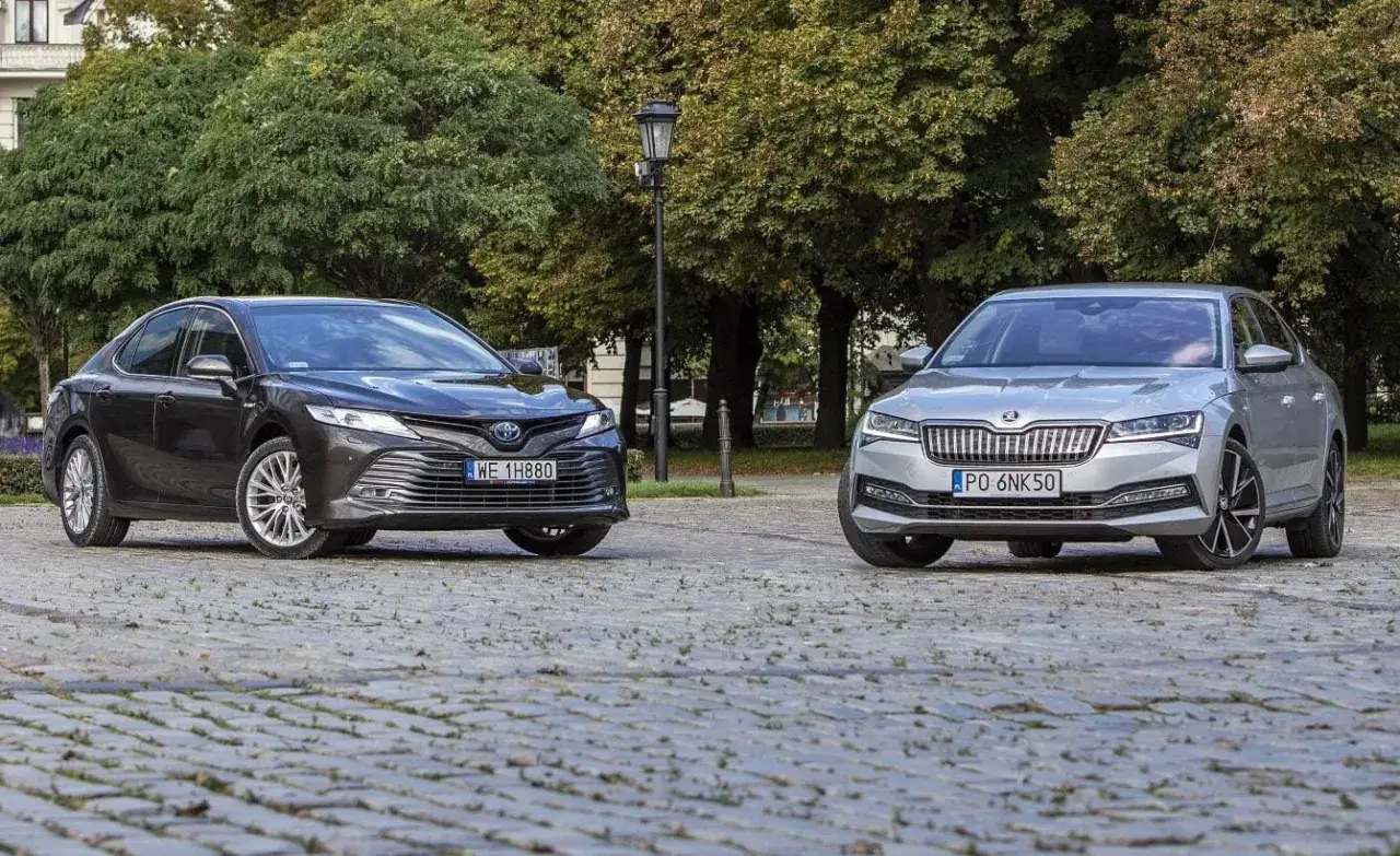 Dwa sedany: ciemny Toyota Camry 2025 i srebrny Skoda Superb, stoją obok siebie na brukowanym placu.