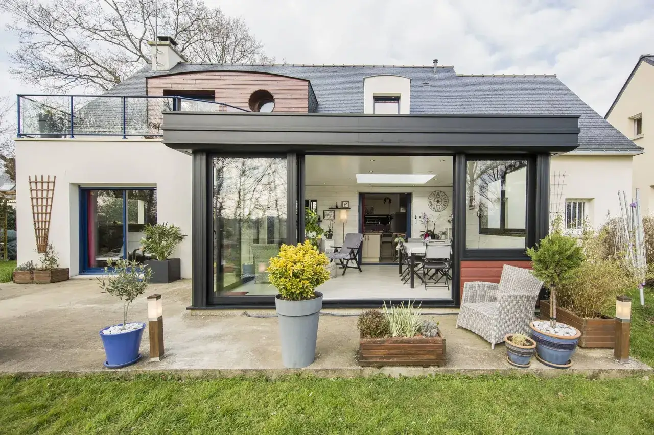 Ampliamento casa con estensione esterna moderna, con ampie vetrate che si aprono su un patio con piante e arredi da esterno.