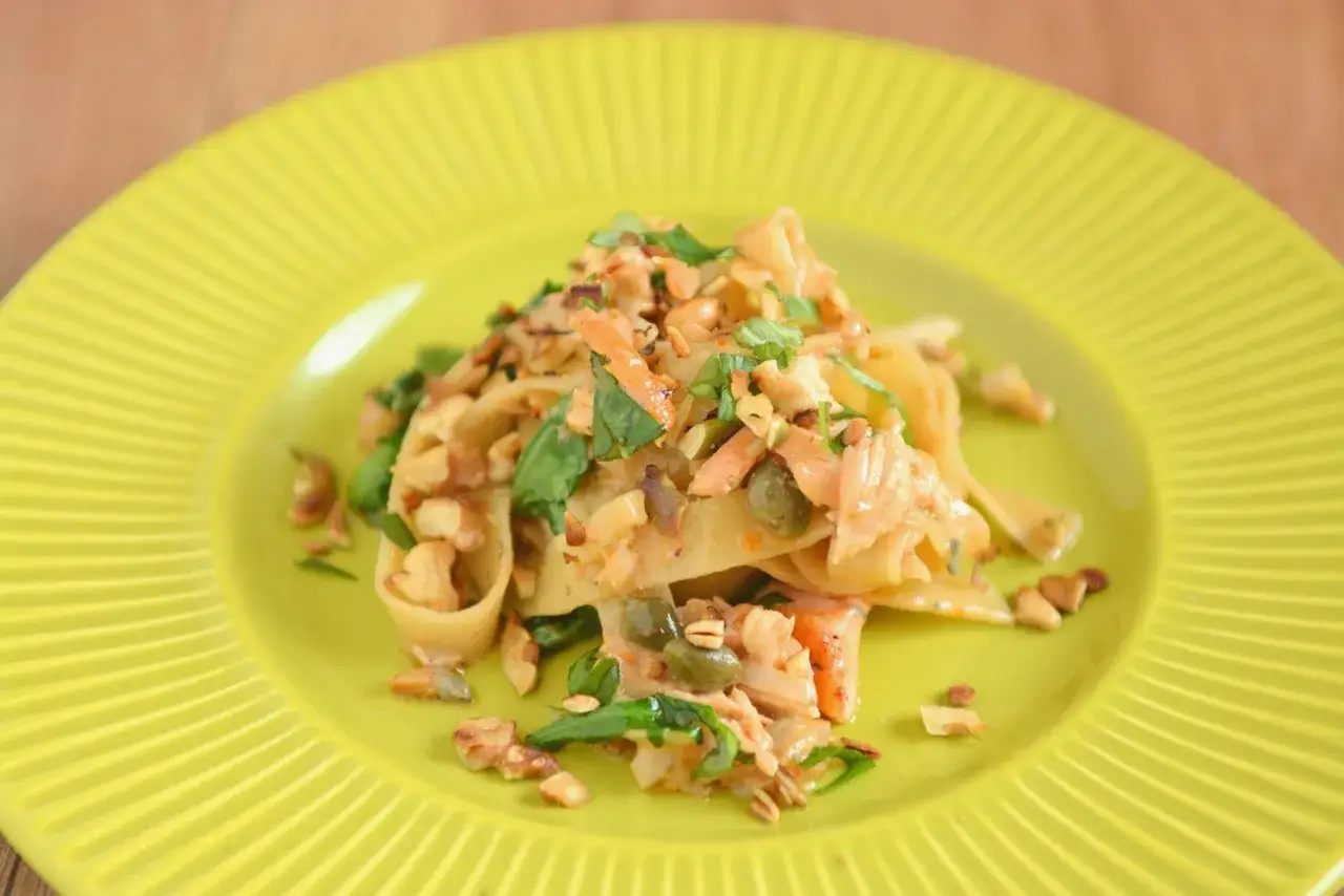 Pappardelle z kawałkami łososia, kaparami i orzechami na żółtym talerzu.