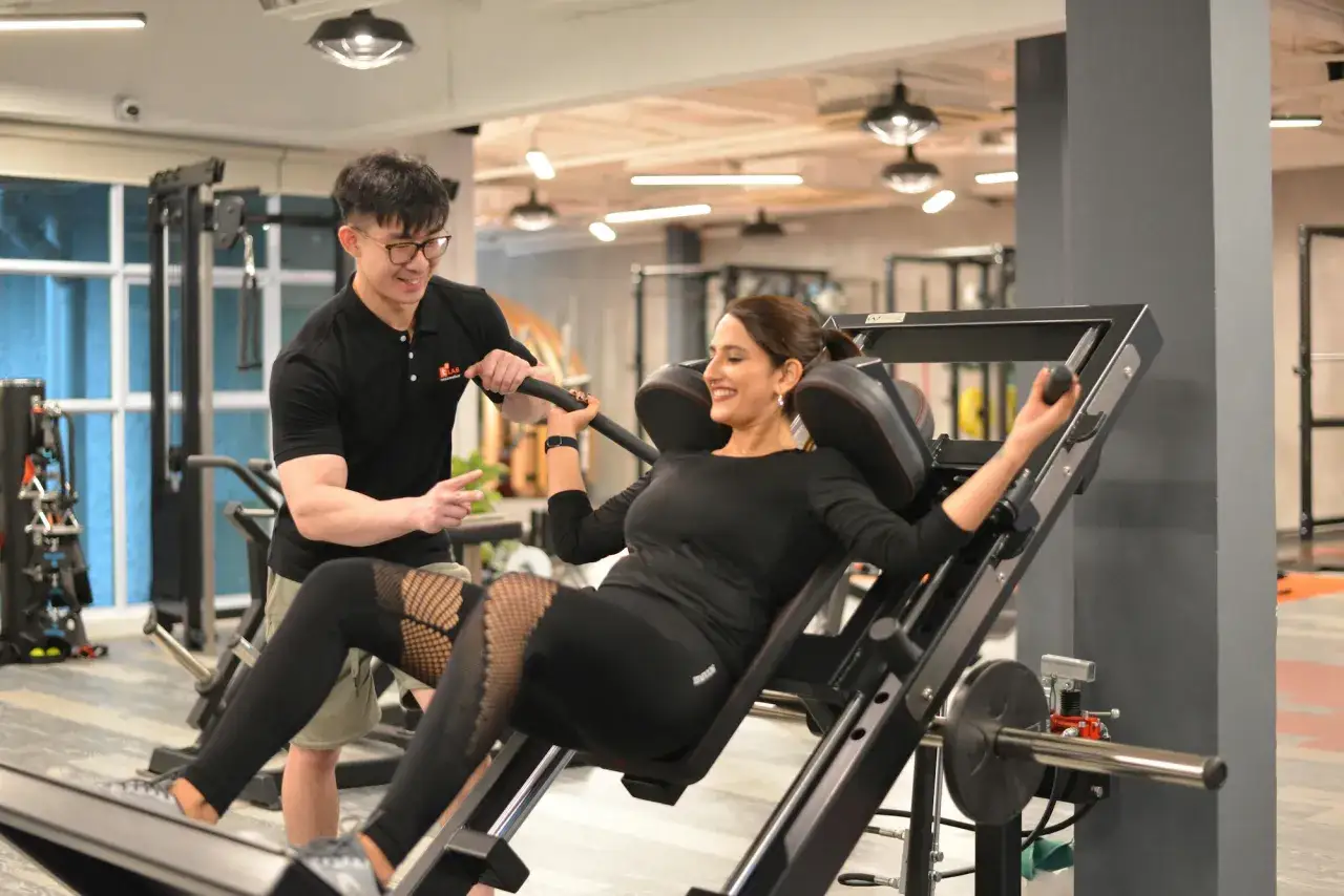 Trener pokazuje kobiecie, od czego zacząć trening na siłowni, asystując przy maszynie do ćwiczeń nóg.