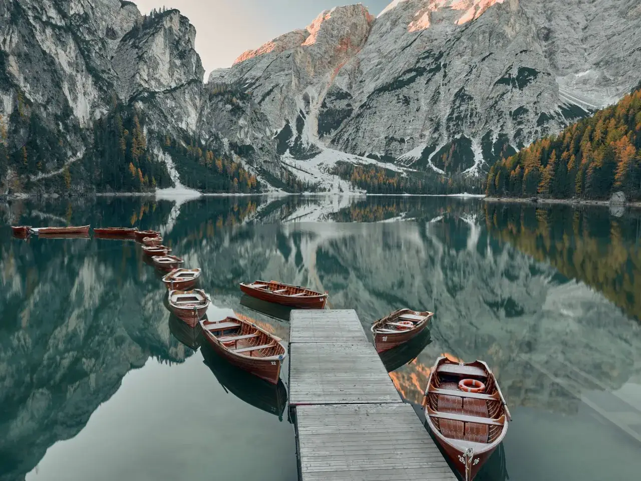 Drewniane łodzie przy drewnianym pomoście na spokojnym jeziorze Lago di Braies, odbijające majestatyczne góry i jesienne drzewa.