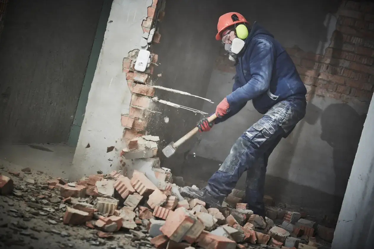 Obrero con casco y mascarilla rompe un tabique de ladrillo con un mazo. El tiempo que se tarda en tirar un tabique depende de la habilidad y la fuerza.