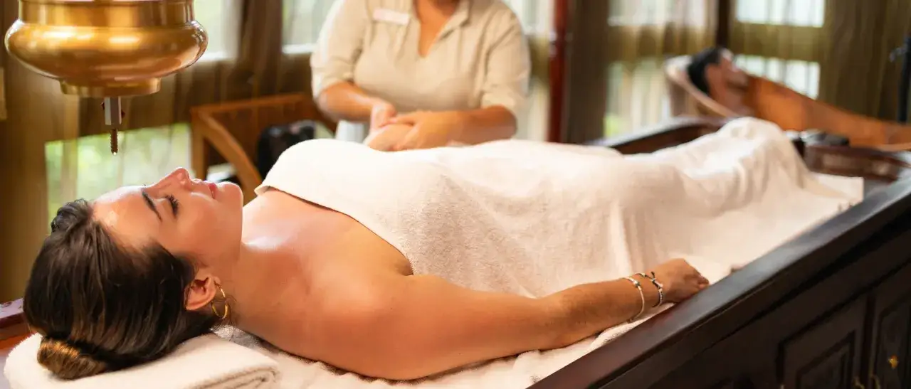 Relaks w spa: kobieta leży na stole do masażu, a nad jej czołem wisi miedziane naczynie, z którego kapie olejek. To kwintesencja tego, co to jest spa.