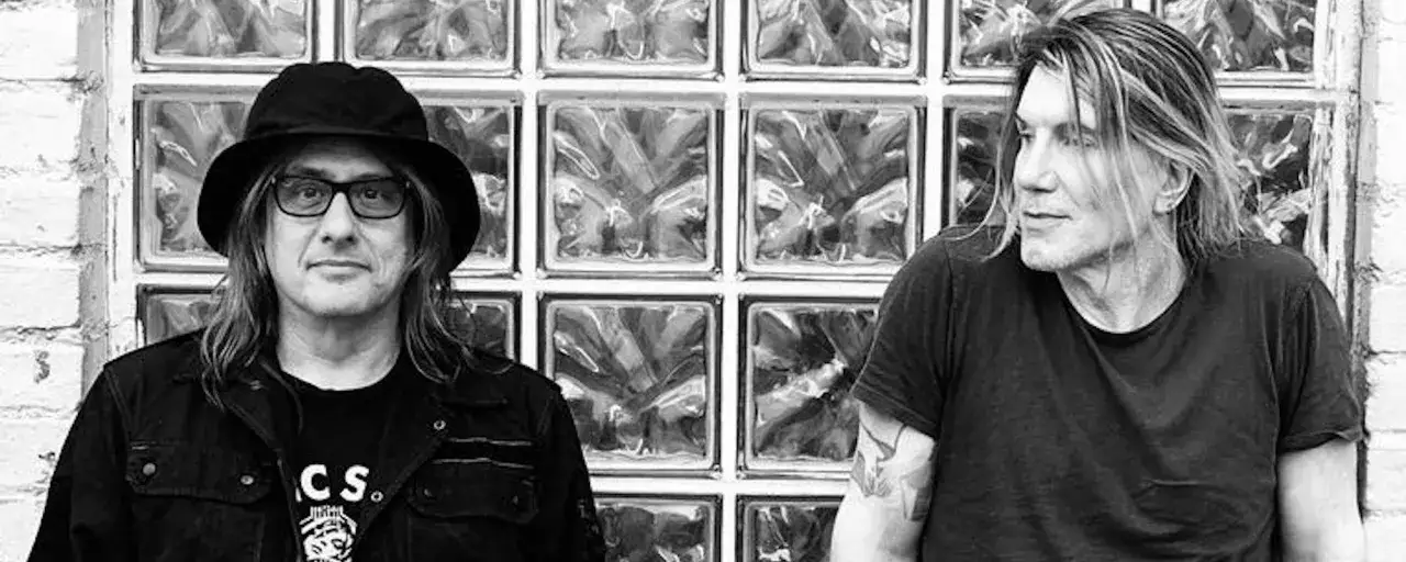 Goo Goo Dolls Iris tekst. Dwóch muzyków zespołu Goo Goo Dolls pozuje przed ścianą z cegły i szkła.