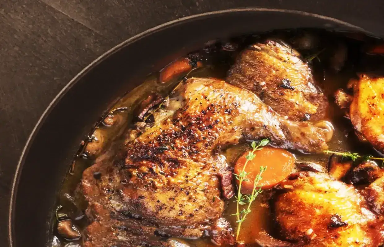 Un plat de coq au vin mijoté dans une cocotte, avec des morceaux de poulet dorés, des carottes, des champignons et des herbes.