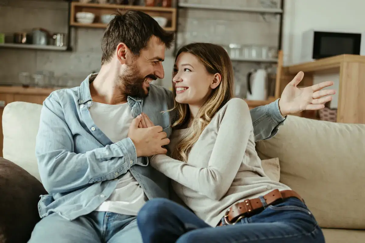 Pareja sonriente en un sof&aacute;, compartiendo un momento &iacute;ntimo. Ella parece explicarle algo, &eacute;l la escucha atentamente, demostrando que el amor puede ser profundo y conectar almas, como en la experiencia demisexual que es.