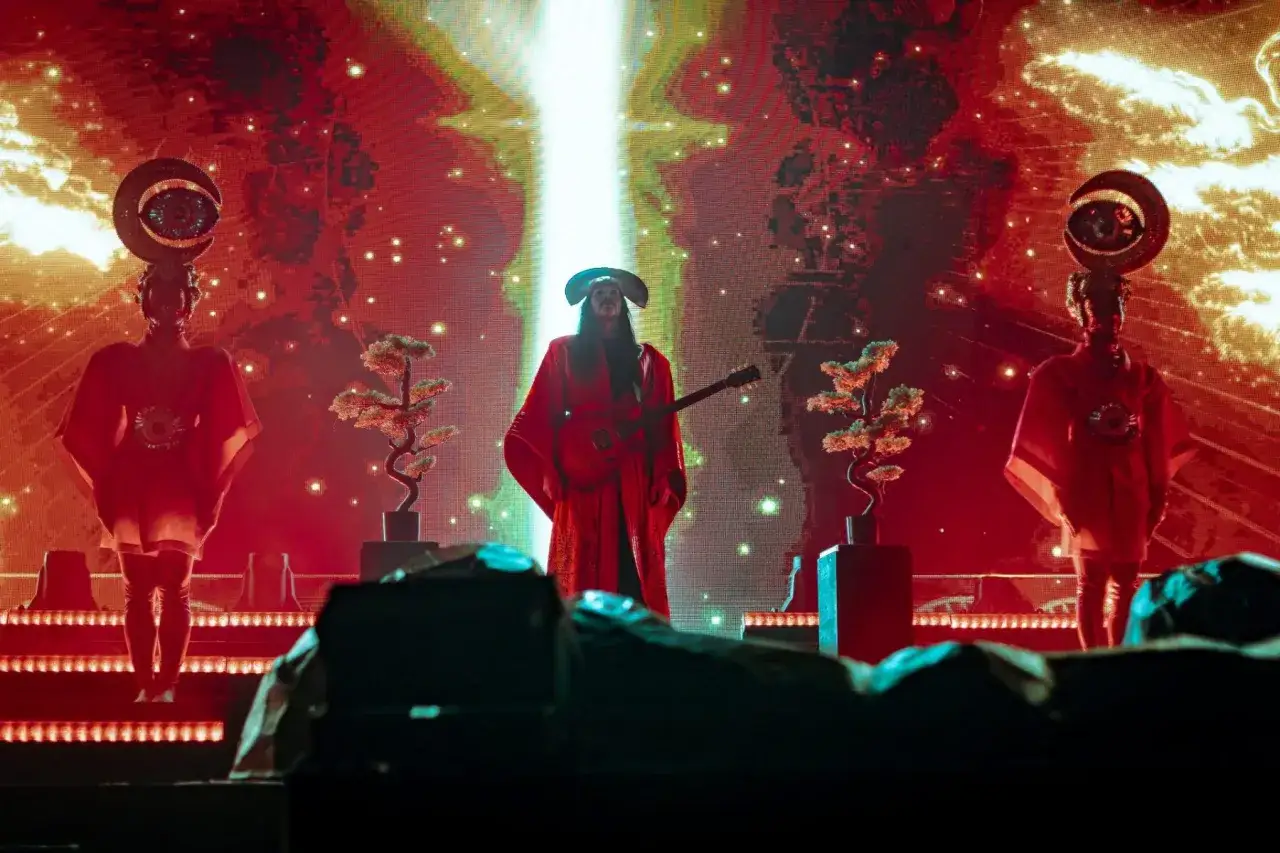 Na scenie, w blasku czerwieni i złota, artysta w stroju przypominającym **empire of the sun** gra na gitarze. Po bokach stoją dwie postacie w czerwonych szatach z ozdobami na głowach.