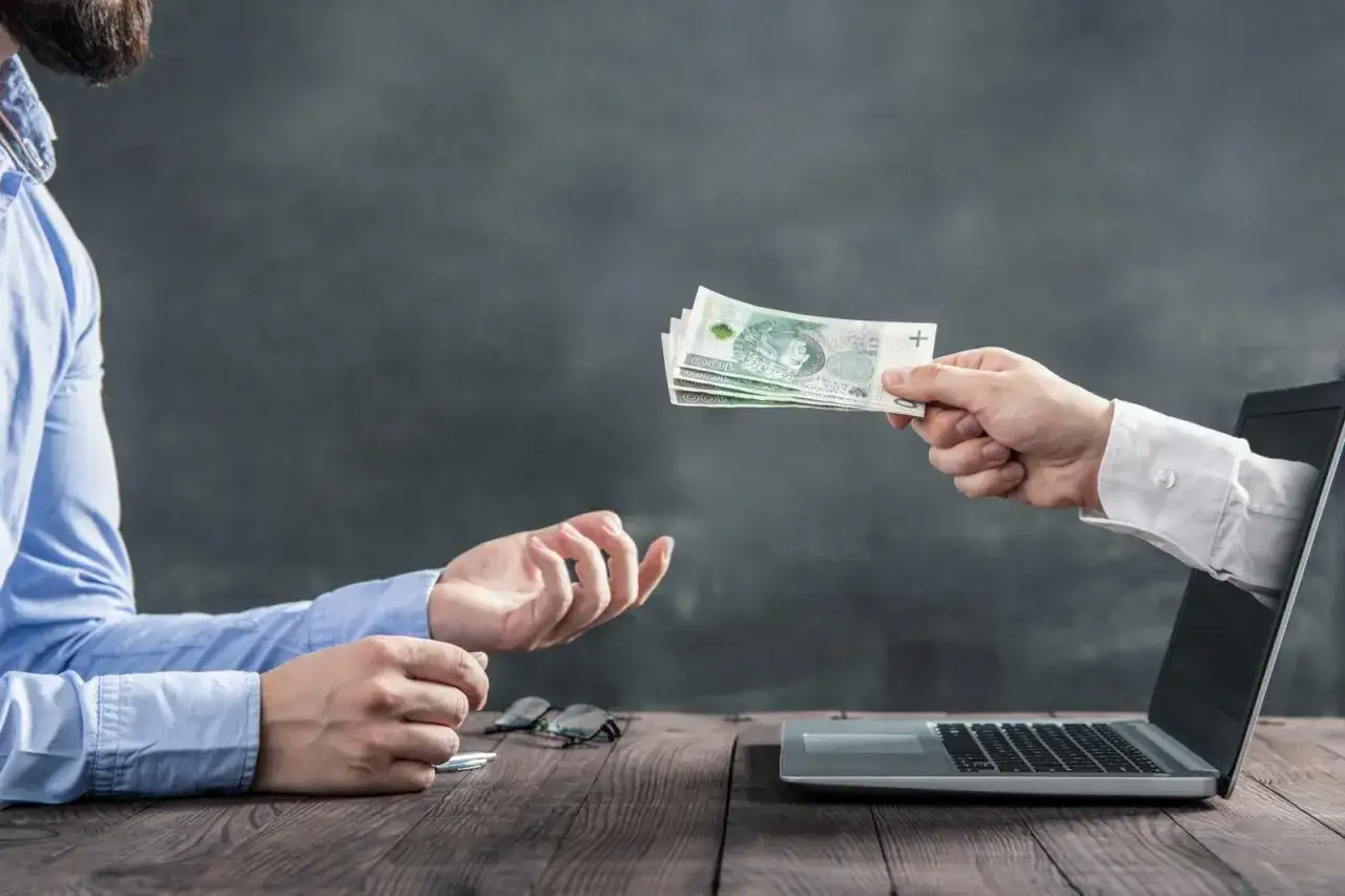 Ręka wychodząca z laptopa podaje banknoty, symbolizując dziedziczenie pieniędzy na rachunku bankowym.