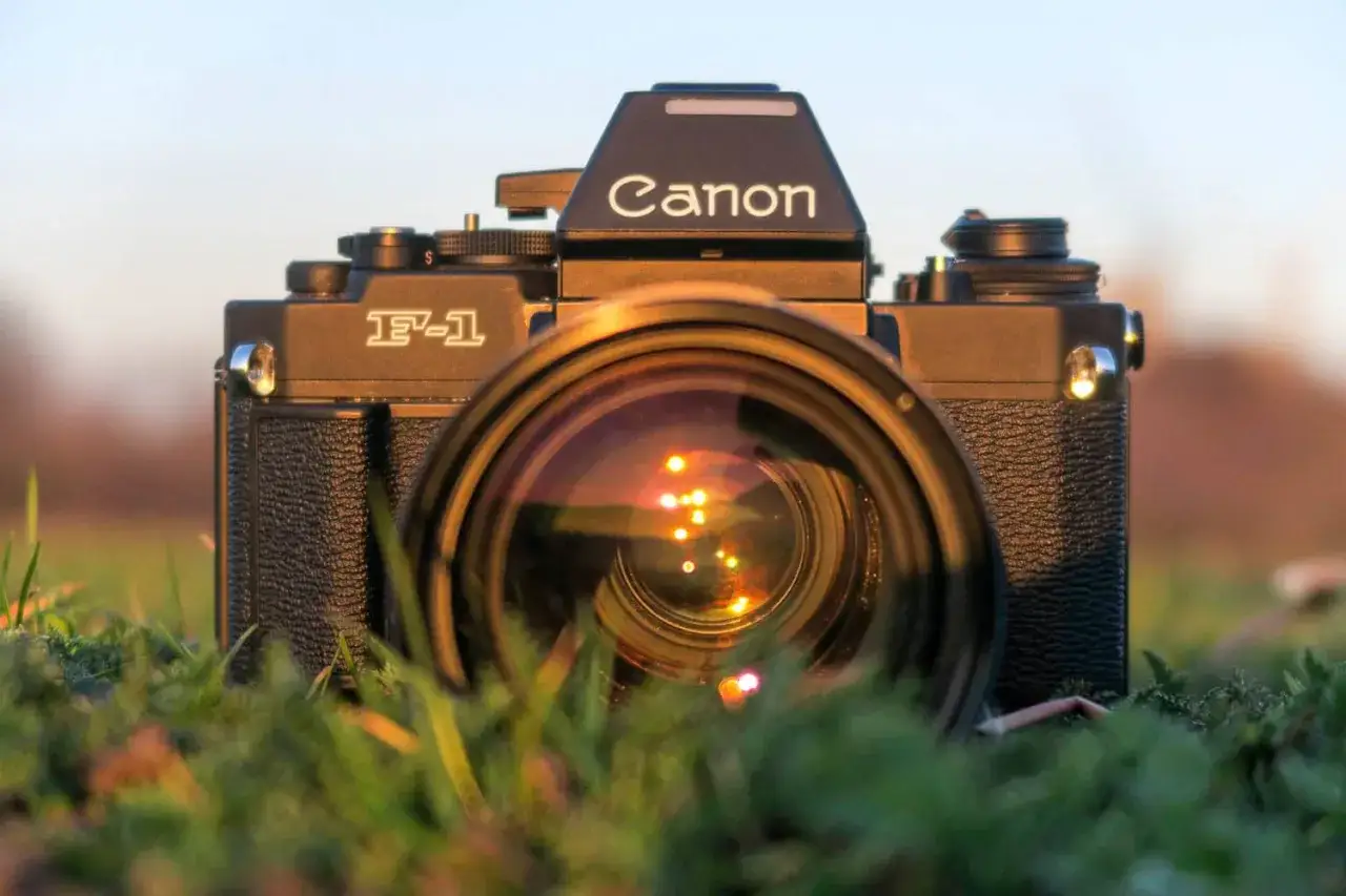 En svart Canon F-1 kamera med objektivet fram&aring;t, placerad i gr&auml;set. Solnedg&aring;ngens reflektioner syns i linsen.