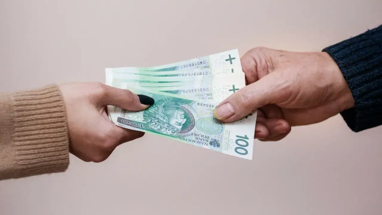 Dwie dłonie przekazują sobie banknoty 100 zł. Może to być symbol zapłaty lub decyzji o ppk rezygnacja.