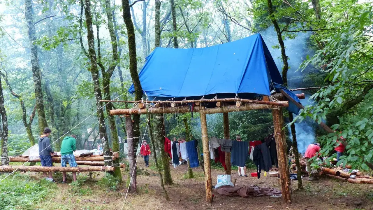 Camp de scout dans la forêt. Une tente bleue est installée sur une structure en bois. Du linge sèche sur une corde. De la fumée s'échappe d'un feu.