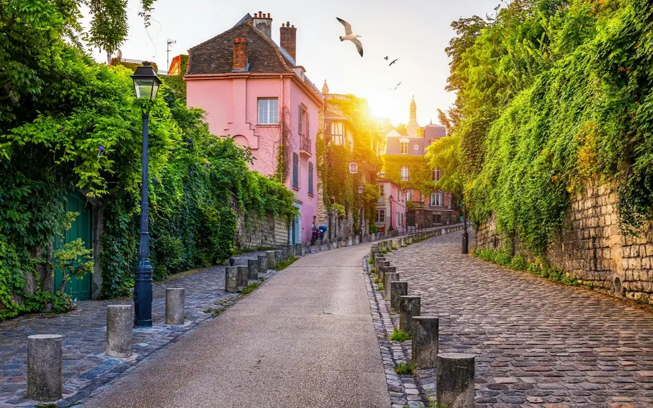 Urokliwa uliczka na Montmartre, z różowymi domami porośniętymi bluszczem i słońcem przebijającym się przez liście.