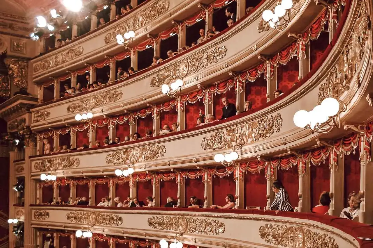Widownia La Scala, złote zdobienia, czerwone kotary i widzowie czekający na rozpoczęcie spektaklu z repertuaru.