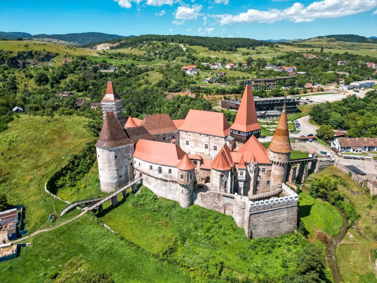Majestatyczny zamek w Transylwanii, otoczony zielonymi wzgórzami i lasami, z charakterystycznymi wieżami i czerwonymi dachami.