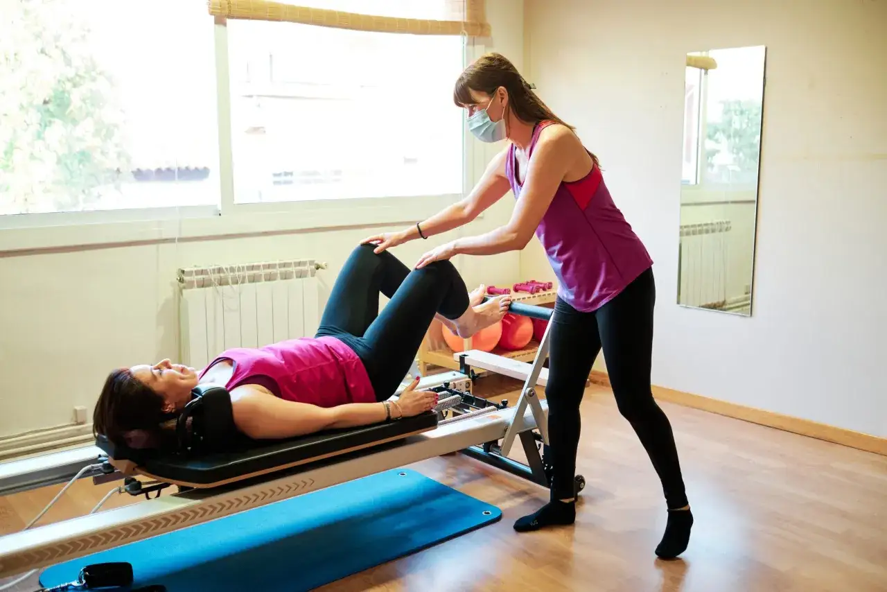 Instruktorka w maseczce pomaga kobiecie na reformerze. To przykład ćwiczeń, które pokazują, co to pilates – budowanie siły i elastyczności.