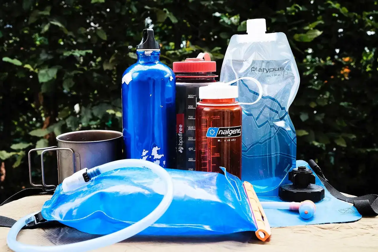 Mehrere Trinkflaschen und ein Trinkbeutel liegen bereit. Eine blaue Trinkflasche mit Erinnerungsfunktion steht neben einer braunen Nalgene-Flasche.
