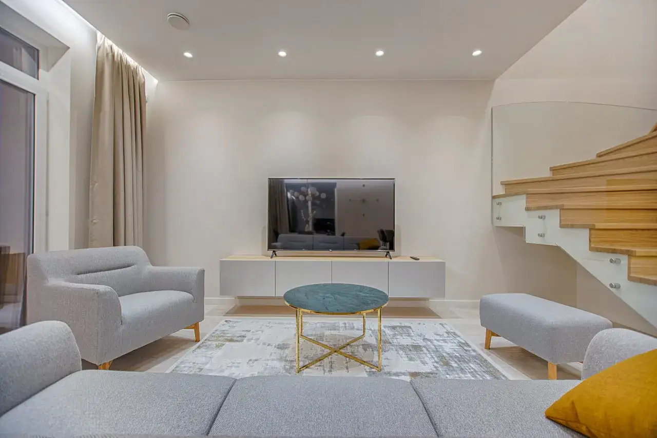 Nowoczesny salon w stylu minimalizm. Szara kanapa, fotel, stolik kawowy i dywan. Drewniane schody z przeszkloną balustradą.