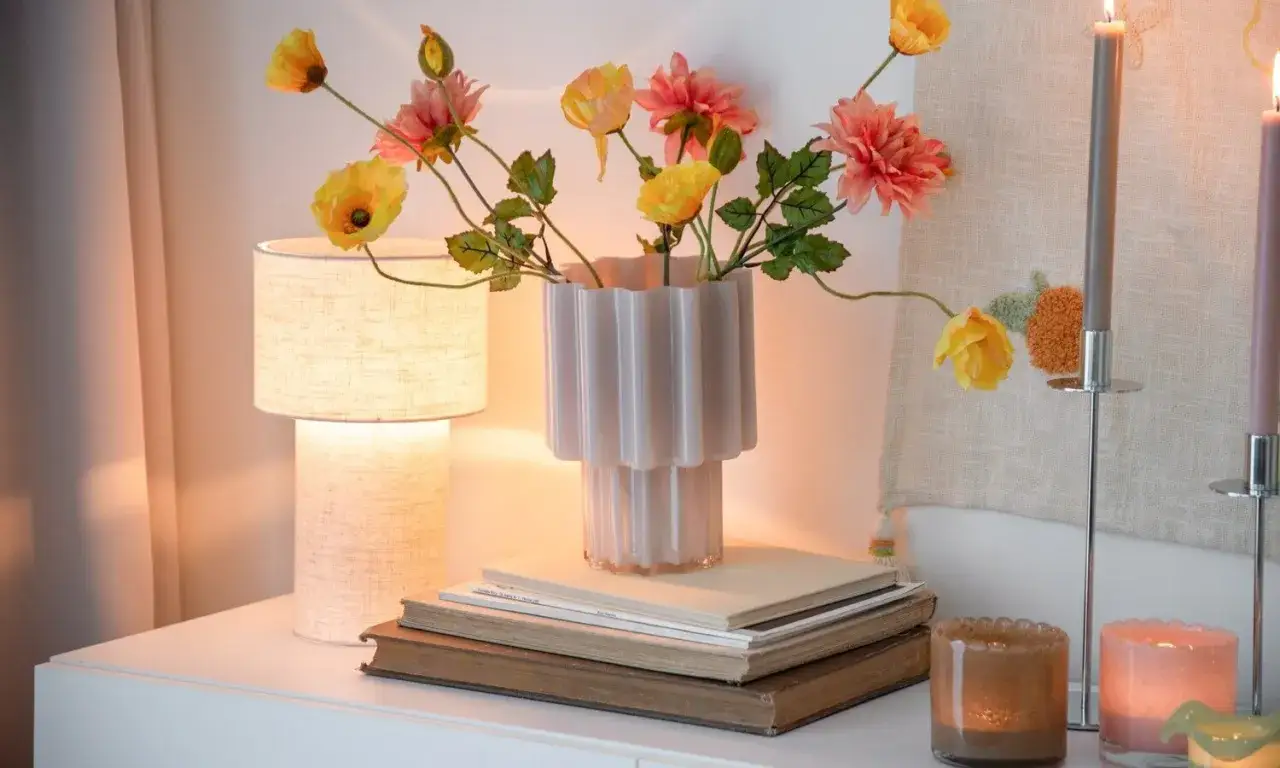 En vas med blommor st&aring;r p&aring; b&ouml;cker. En lampa ger ett varmt, riktat ljus. Ljus och dekorationer skapar en mysig st&auml;mning.