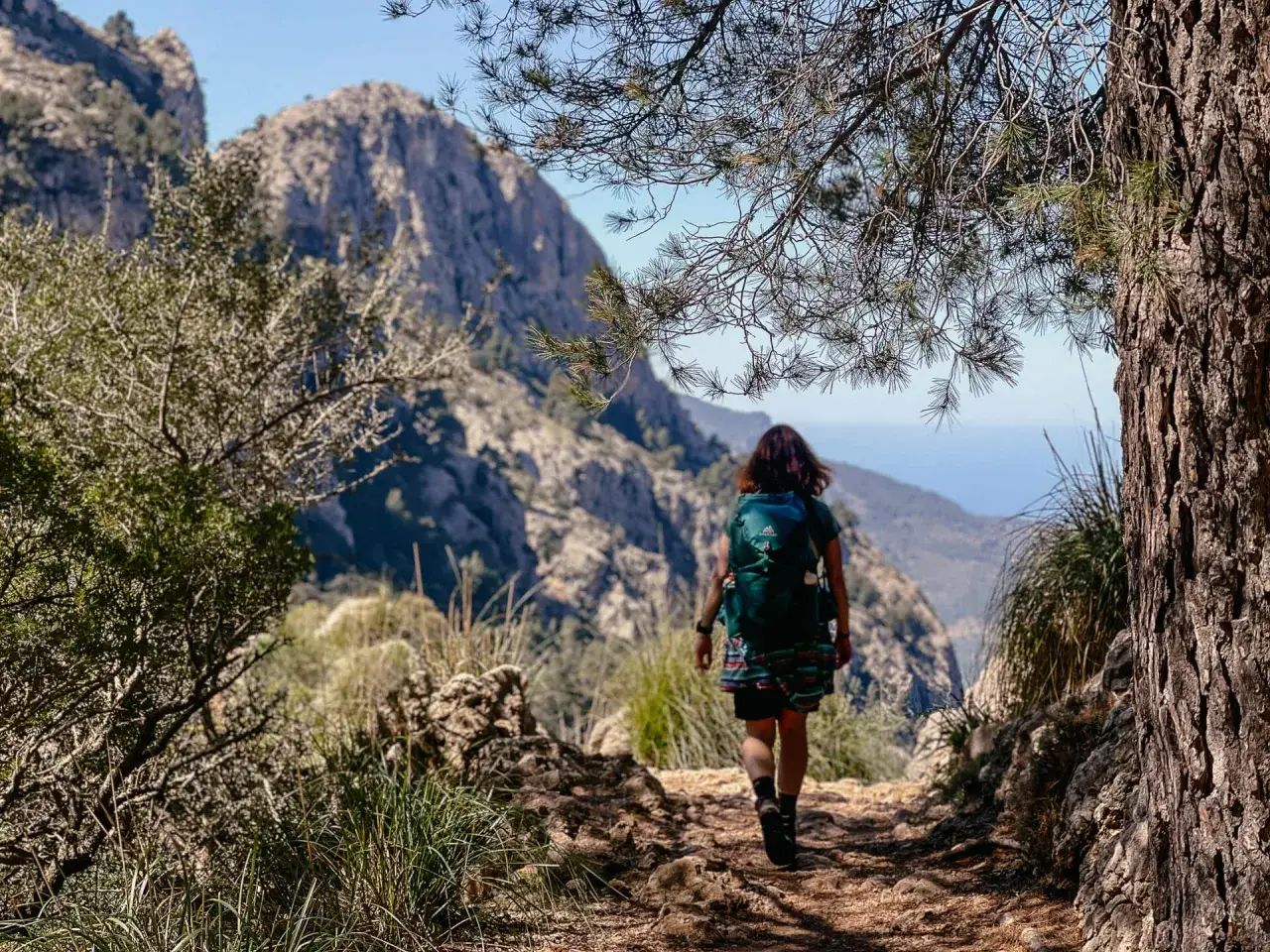 Eine Person mit Rucksack wandert auf einem steinigen Pfad in den Bergen Mallorcas. Im Hintergrund erheben sich felsige Gipfel und das blaue Meer.