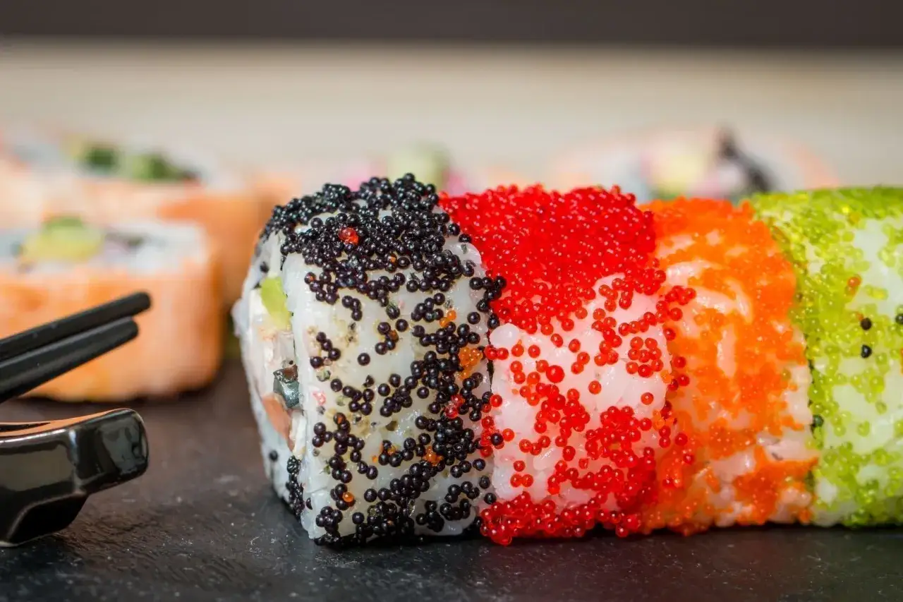 Rolki sushi z czarnym, czerwonym, pomarańczowym i zielonym tobiko. Wyglądają apetycznie obok pałeczek.
