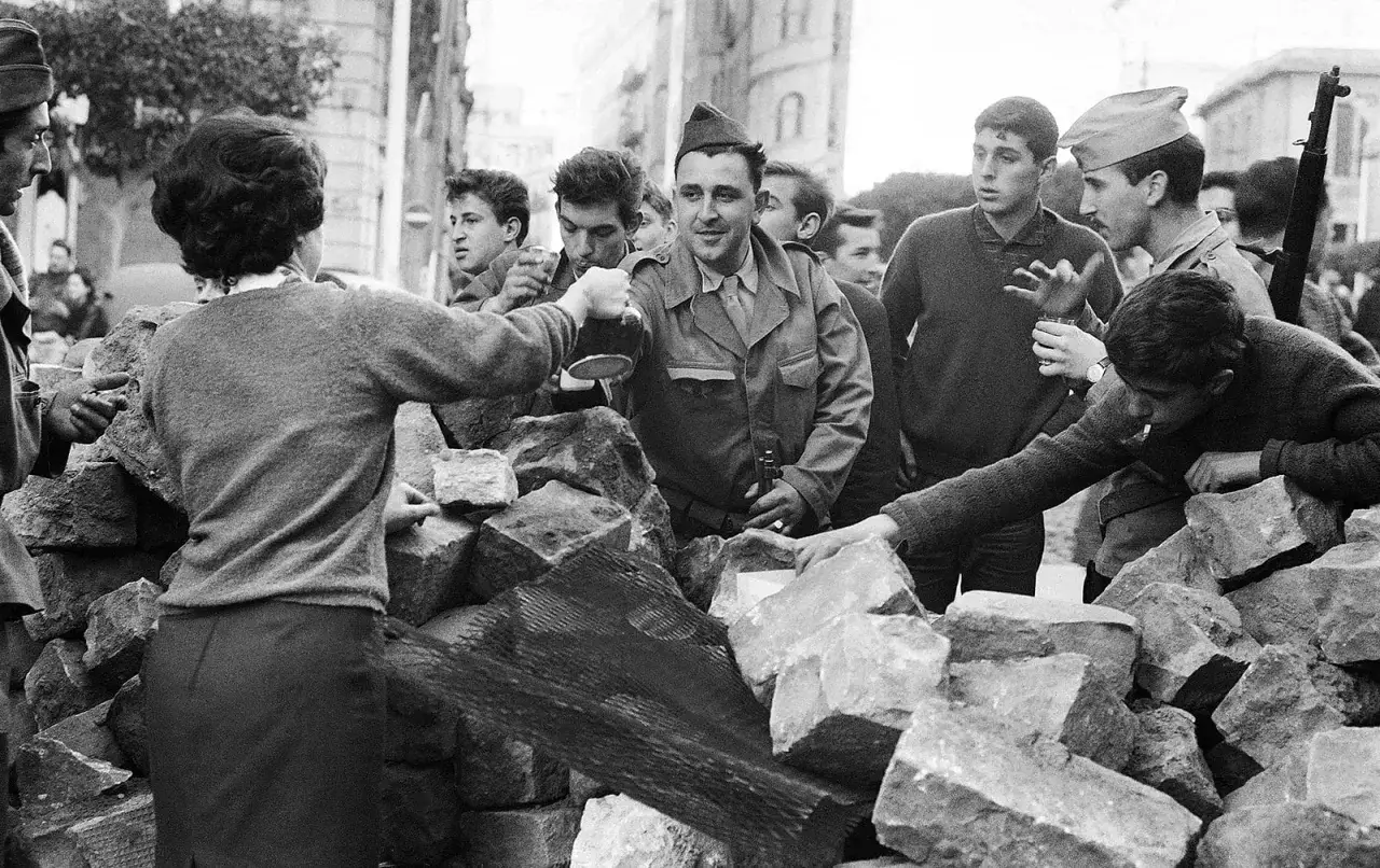 Un grupo de j&oacute;venes, algunos con uniformes militares, interact&uacute;an tras una barricada de piedras en Argelia. Un hombre sirve bebida, mientras otros observan o fuman.