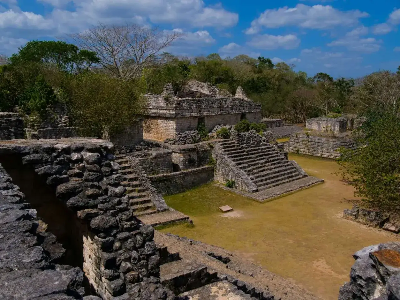 Ruinen einer alten aztekischen Pyramide, umgeben von &uuml;ppiger Vegetation und unter einem blauen Himmel mit Wolken.