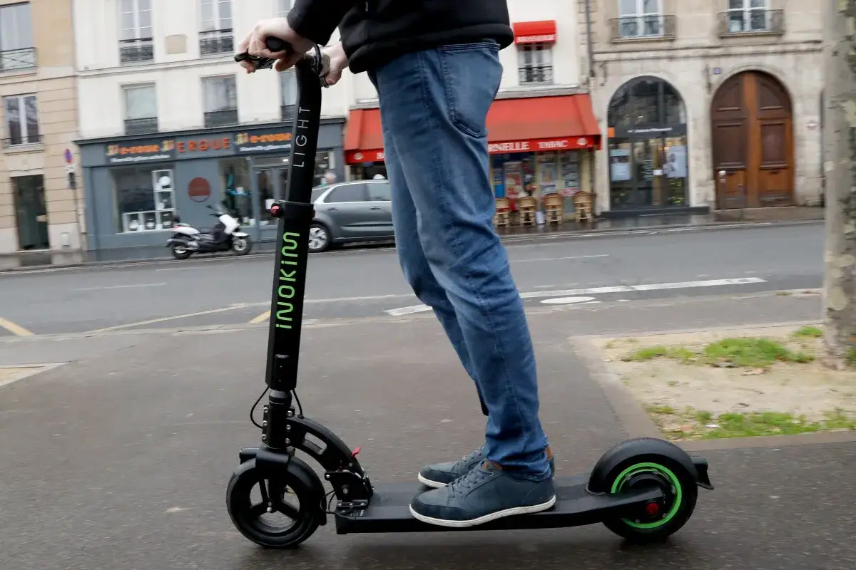 Un homme roule en trottinette électrique sur un trottoir, respectant la loi trottinette électrique. La ville est en arrière-plan.