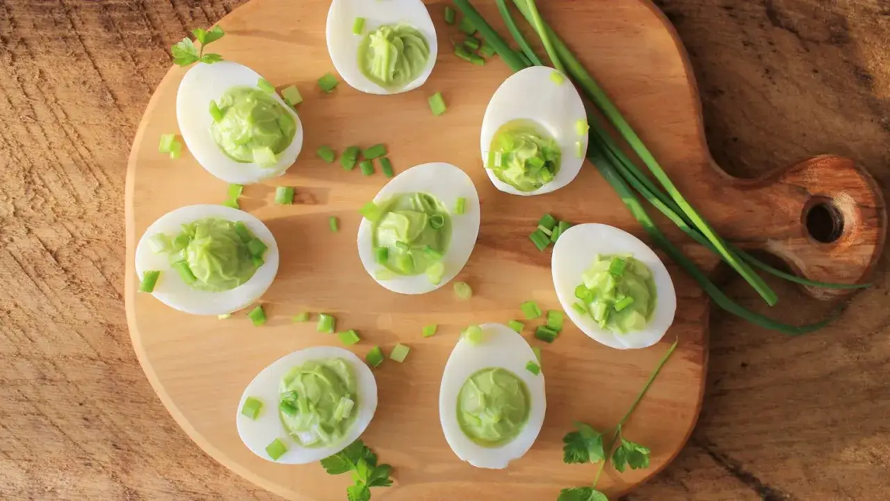 Jajka faszerowane guacamole, ozdobione szczypiorkiem i pietruszką, prezentują się apetycznie na drewnianej desce.