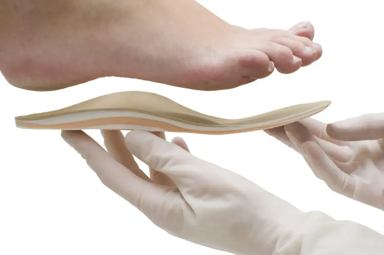Un pied au-dessus d'une semelle orthop&eacute;dique tenue par des mains gant&eacute;es. Une podologue ordonnance ou pas, le soin adapt&eacute;.